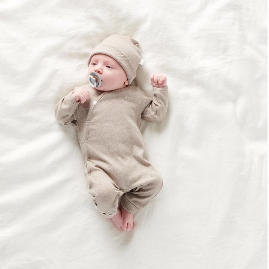 Baby liggend op een witte achtergrond, draagt een beige boxpakje en muts.
