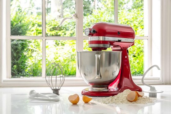 Robot pâtissier KitchenAid Artisan rouge Empire avec bol argenté, fouet, crochet pétrisseur et feuille sur un comptoir.