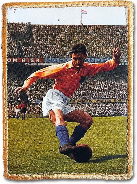 Voetballer in oranje shirt, witte broek en blauwe sokken, met bal op grasveld voor publiek.