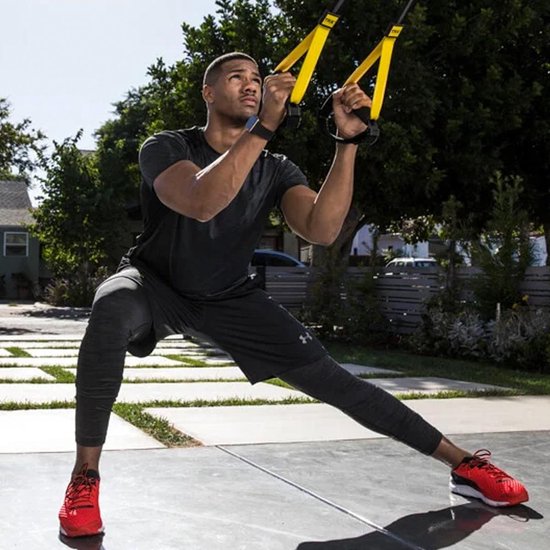 Man met donker haar en rode schoenen doet een squat met TRX-banden buiten.