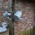 Drie zinken gieters hangen aan een houten rek tegen een bakstenen muur. Twee kleine gieters liggen op een ton.