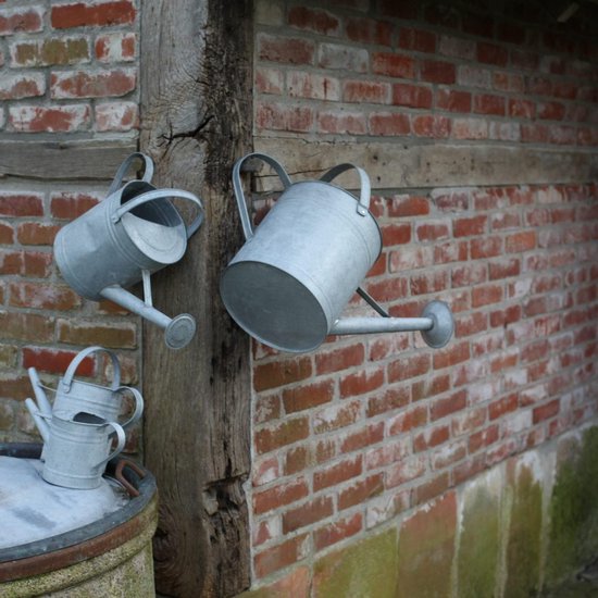 Drie zinken gieters hangen aan een houten rek tegen een bakstenen muur. Twee kleine gieters liggen op een ton.