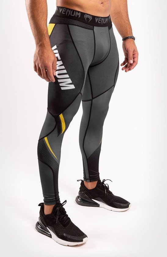 Man draagt een grijze compressietight met gele accenten, "Venum" logo op het been en zwarte mesh.