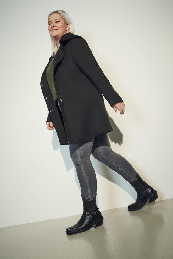 Model met zwart jack met capuchon, groen shirt en grijze jeans.