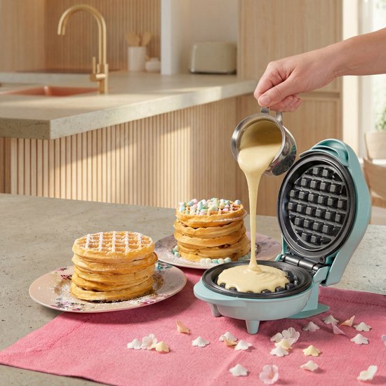 Blauw wafelijzer in keuken. Beslag wordt gegoten in het wafelijzer. Twee stapels wafels met fruit en chocolade ernaast.