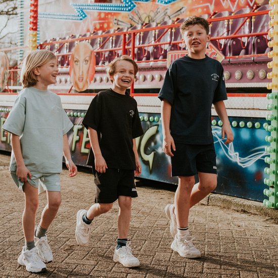 Drie lachende jongens lopen op een bestrate ondergrond. Ze dragen bijpassende t-shirts en shorts in lichtblauw, zwart en donkerblauw.