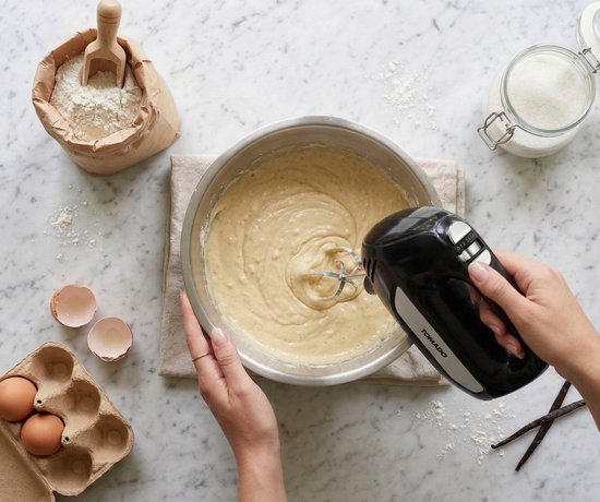 Zwarte Tomado handmixer met garde in een kom met beslag, naast eieren, bloem en suiker.
