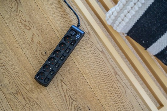 Zwarte stekkerdoos met 6 stopcontacten en individuele schakelaars, liggend op een houten vloer.