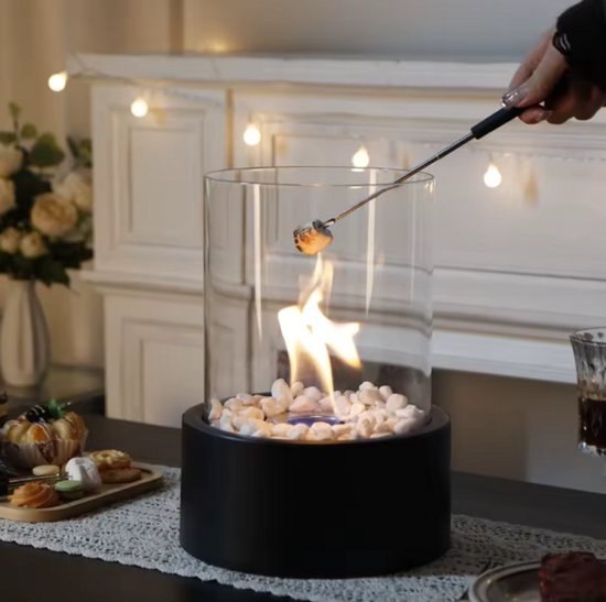 Cilindrische tafelhaard met zwarte basis, glazen omhulsel en witte stenen, met een vlam en een marshmallow aan een spies.