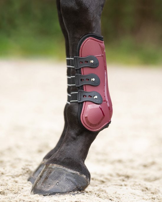 Donkerrode peesbeschermer om een paardenbeen met glanzende voorkant en zwarte sluitingen.