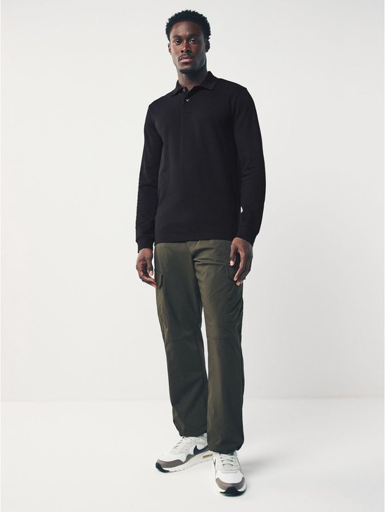 Man, staand, draagt een zwart piqué poloshirt met lange mouwen en legergroene cargo broek, witte sneakers.