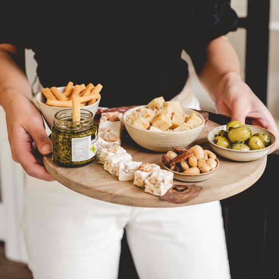 Vrouw met witte broek houdt een houten plank vast met olijven, noten, kaasblokjes, nougat, pesto, grissini en plakjes worst.
