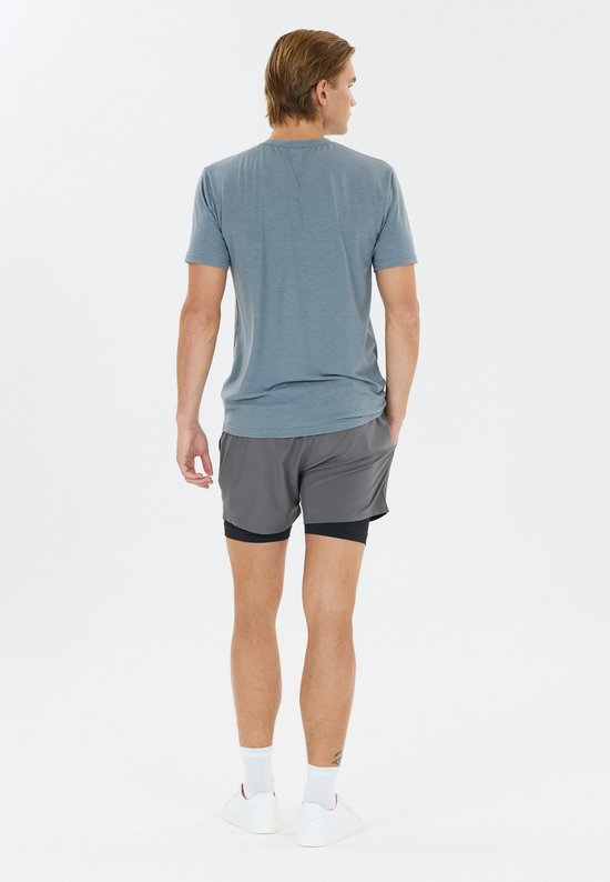 Van de achterkant gezien, man in blauw shirt en grijze sportshort met zwart binnenbroekje, witte sokken en witte schoenen.