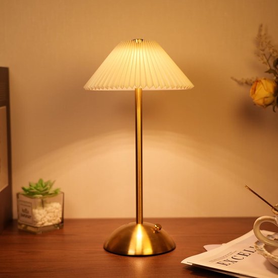 Een gouden tafellamp met geplooide kap staat op een bureau naast een plant en een boek.