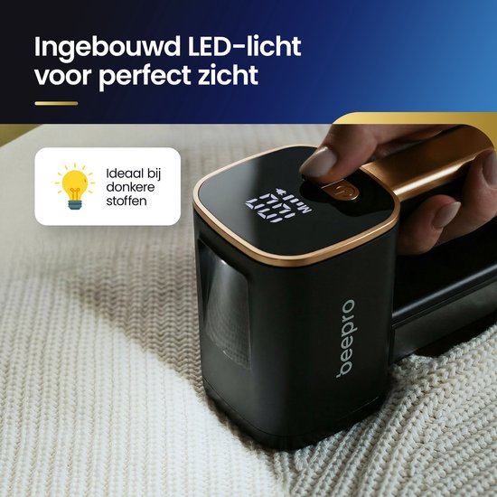 Een hand houdt een zwarte en gouden pluizenverwijderaar vast met een ingebouwd LED-licht, die op een witte gebreide stof wordt gebruikt. Het display toont de instellingen.