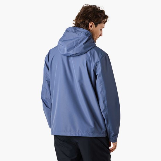 Man kijkt naar achteren, draagt een blauw X-Trail windjack met capuchon en zwarte broek.
