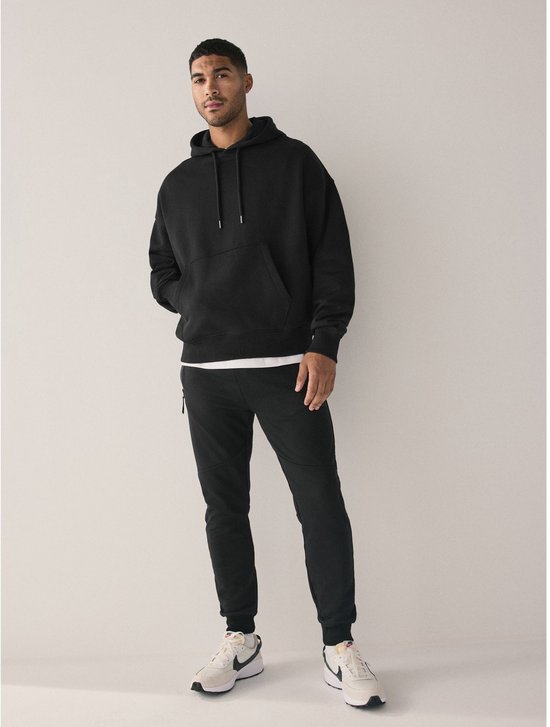 Man draagt een zwarte oversized hoodie met capuchon, een witte T-shirt, zwarte broek en witte sneakers.