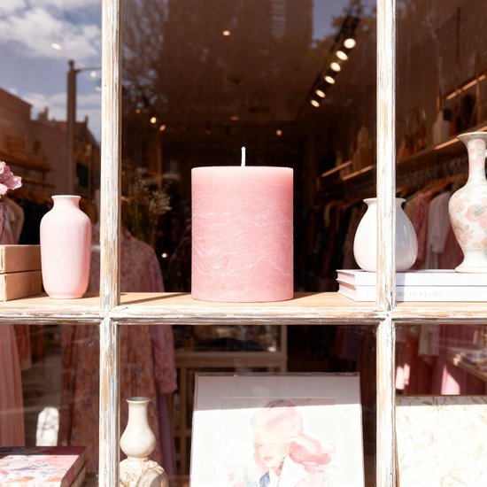 Roze stompkaars met decoratieve textuur in etalage, tussen vazen en kledingrek.