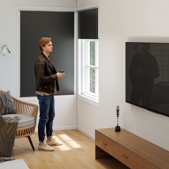 Man in leren jas en spijkerbroek staat bij een raam met donkergrijs rolgordijn. Een stoel en tv zijn zichtbaar.