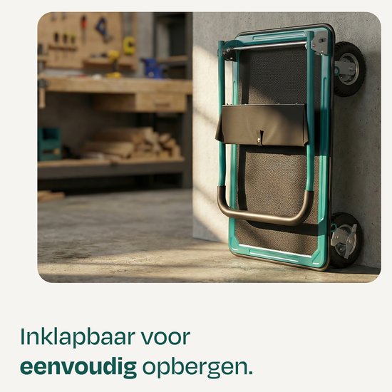 Ingeklapte groene plateauwagen leunend tegen een betonnen muur, met werkplaats in de achtergrond. Geschikt voor eenvoudige opslag.