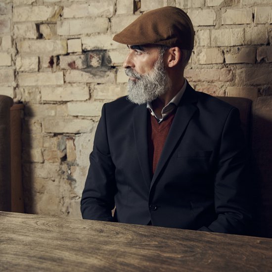 Bruine pet op een man met grijze baard en zwart jasje aan tafel.