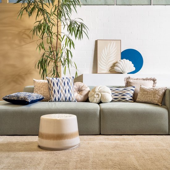 Een woonkamer met een bank vol sierkussens, waaronder een wit bouclé kussen in schelpvorm.