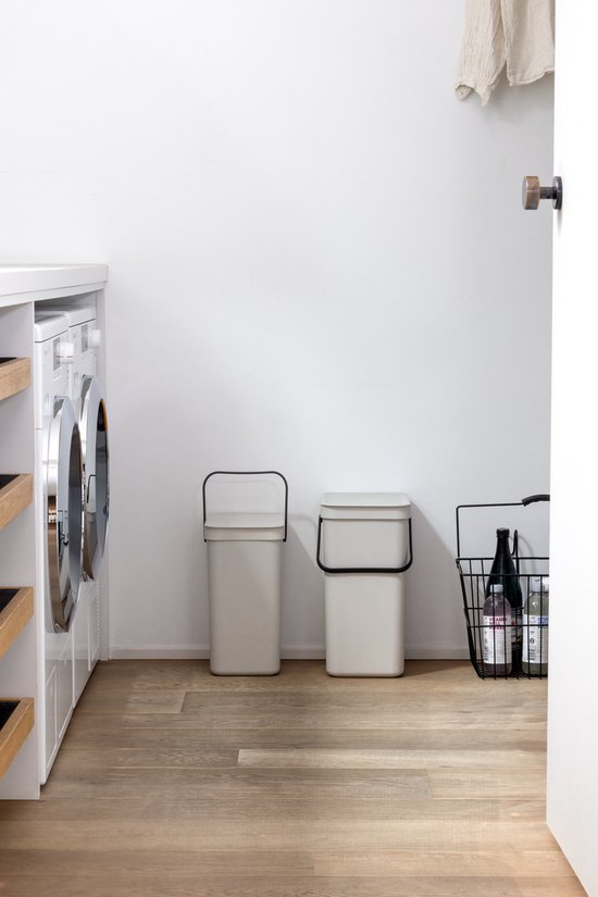 Twee licht beige prullenbakken met zwarte handvatten naast een wasmachine.