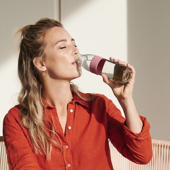 Vrouw met blond haar in oranje blouse drinkt uit een Mepal waterfles met roze band.