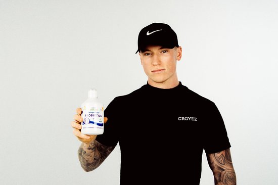 Jonge man met tatoeages, draagt een zwarte pet en een zwart T-shirt, houdt een flesje PROBIOTICS omhoog.