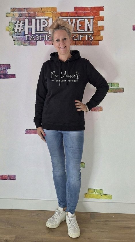 Zwarte hoodie met de tekst "Be Yourself and don't Apologize". Gedragen door een vrouw met een spijkerbroek en witte sneakers.