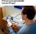 Man met blauwe trui gebruikt zijn smartphone om de HP app te bedienen, terwijl hij naast een witte HP printer staat.