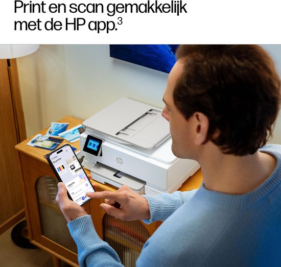 Man met blauwe trui gebruikt zijn smartphone om de HP app te bedienen, terwijl hij naast een witte HP printer staat.