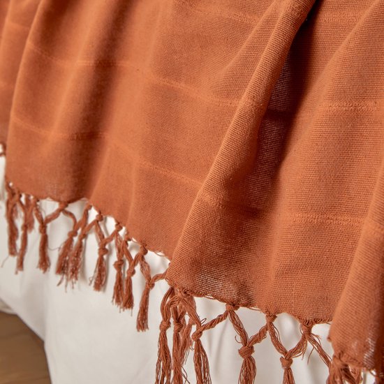 Dichtbij op een terracotta plaid met franjes die over een wit bed hangen.