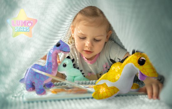 Een klein meisje leest een boek onder een deken, omringd door paarse, lichtgroene en gele dinosaurusknuffels.