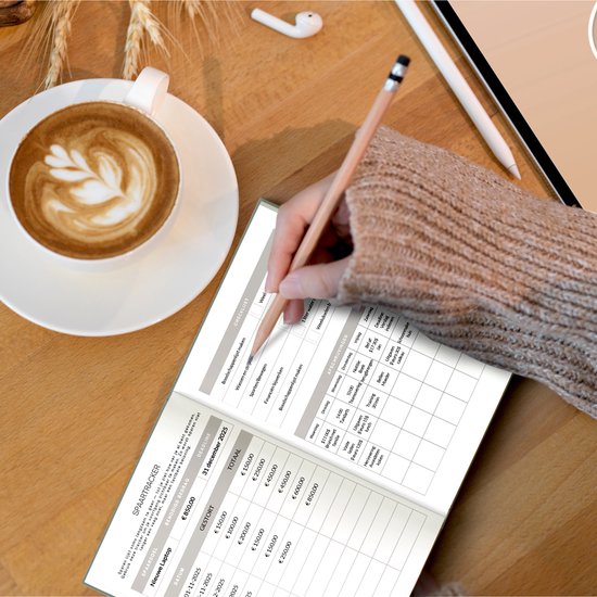 Vrouw met bril schrijft in een budgetplanner. Op tafel een kopje koffie met latte art en een tablet. De planner toont een spaartracker met datum en bedragen.