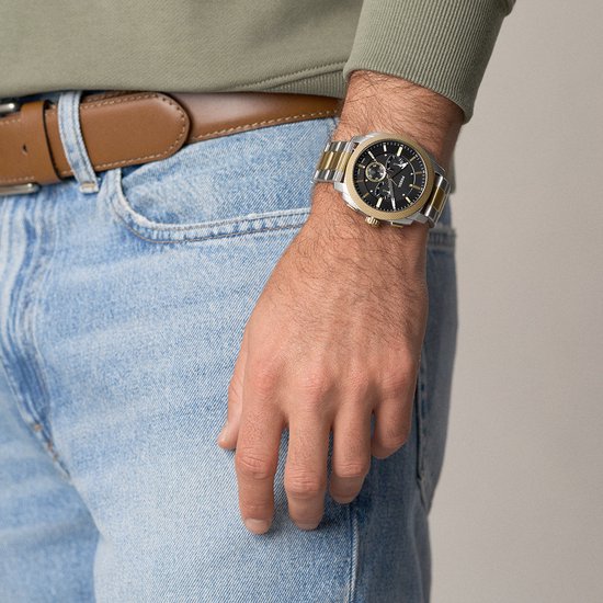Gouden en zilverkleurig Fossil horloge om een mannelijke pols, gedragen met spijkerbroek en bruine riem.