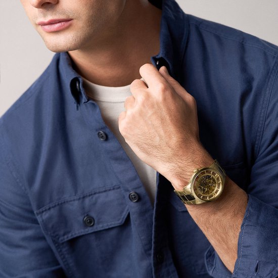 Man met een goudkleurig Fossil skeleton horloge om de pols, gedragen met een blauw overhemd.