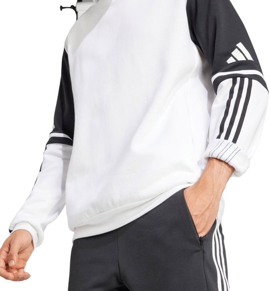 Man in een witte capuchontrui met zwarte mouwen met witte Adidas-strepen. Hij stopt zijn hand in zijn broekzak.