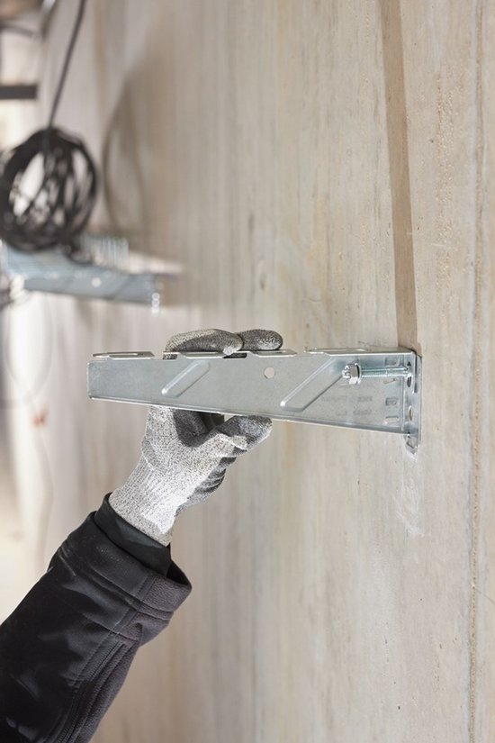 Hand met grijze werkhandschoen bevestigt een metalen anker aan een betonnen muur met een hamer.