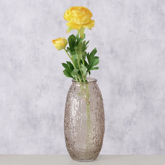 Bruin-beige glazen vaas met een ruw, bobbelig oppervlak, gevuld met een gele bloem en knoppen, tegen een grijze achtergrond.