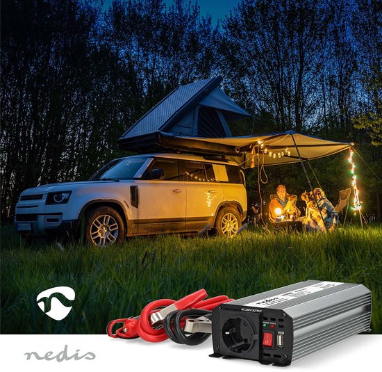 Een zilverkleurige omvormer met rode en zwarte accuklemmen, naast een kampeersetting met een auto, tent en verlichting.