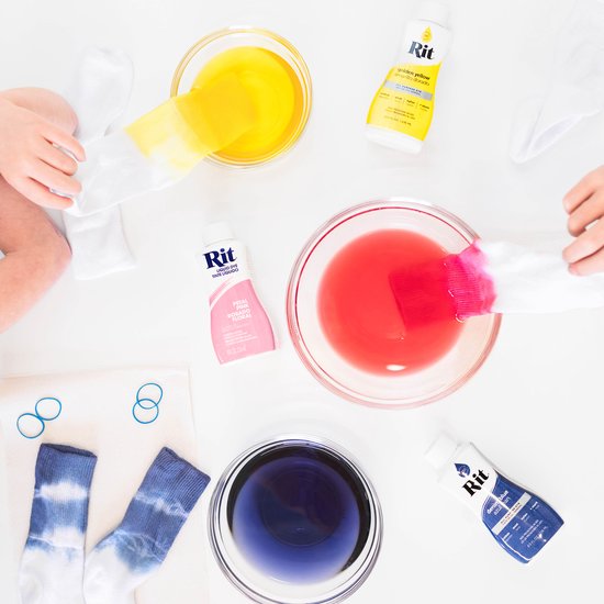 Kinderen verven sokken en handschoenen in kommen met gele, roze en paarse Rit-verf. Naast de kommen liggen flessen verf, elastiekjes en twee geverfde sokken.