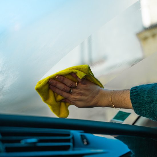 Een gele microvezeldoek veegt een beslagen autoruit schoon. De hand draagt ringen.