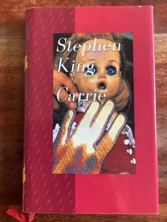 Het boek Carrie van Stephen King met een rode kaft met een afbeelding van een pop en een hand.