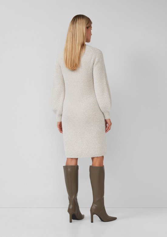 Vrouw met blonde haren, in gebreide jurk met pofmouwen en hoge hals, beige met glitters. Zichtbaar van achteren.