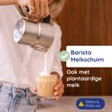 Vrouw giet melkschuim uit zilverkleurige kan in een glas met koffie.