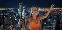 Een lachende vrouw met oranje sporttop, houdt een Insta360 camera op een selfiestick omhoog met een stadsgezicht op de achtergrond.