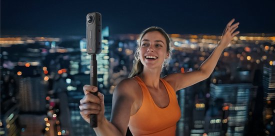 Een lachende vrouw met oranje sporttop, houdt een Insta360 camera op een selfiestick omhoog met een stadsgezicht op de achtergrond.