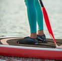 Voeten van een persoon op een paddleboard, dragend zwarte waterschoenen met blauwe stippen en kleurrijke zolen.
