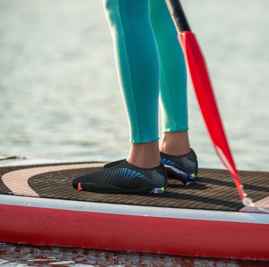 Voeten van een persoon op een paddleboard, dragend zwarte waterschoenen met blauwe stippen en kleurrijke zolen.